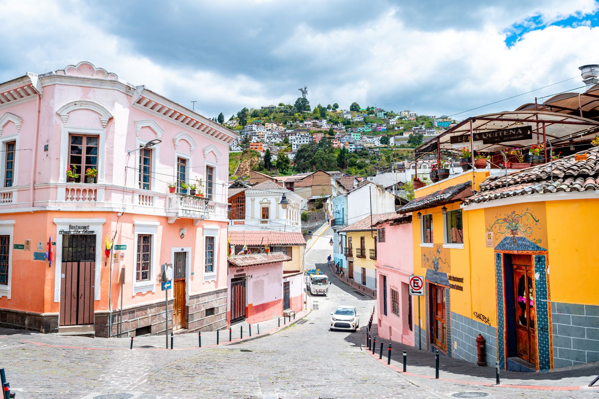 A cobbled street with colorful buildings, other houses on a hill in the background