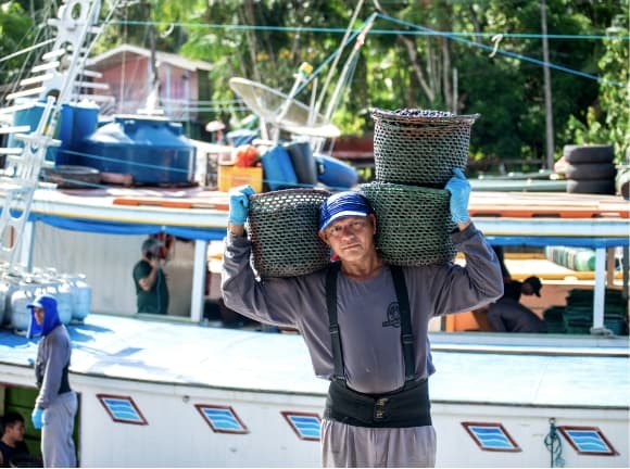 A man transporting baskets of açai on his shoulders, boat in the background